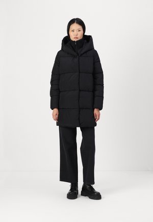 Svart pufferjakke med hette, med quilted design, glidelås foran og lange ermer, kombinert med vide svarte bukser og støvler.