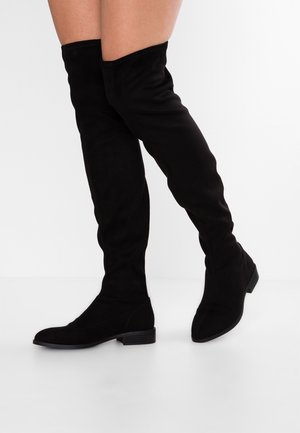 Jambes portant des bottes plates en daim noir au-dessus du genou, debout sur une surface blanche avec un fond blanc uni.