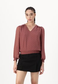 Blusa rosa con tessuto texture, scollo a V, maniche lunghe a sbuffo e orlo arricciato, abbinata a una gonna nera.