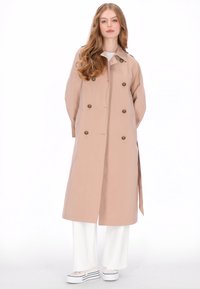 Lichtroze, dubbelgeknoopte trenchcoat met gouden knopen, lange mouwen, zijzakken en een ceintuur in de taille. Draagt over witte broek en schoenen.