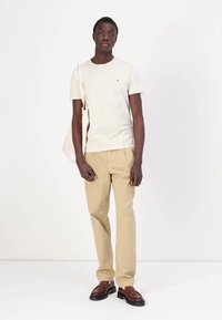 Beige bomull T-skjorte, beige bukser, brune lærersko, holder en lys totebag. Enkel design, nøytrale farger, uformell passform.