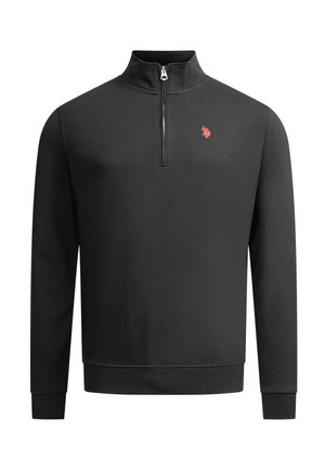Mann steht, trägt einen schwarzen Viertel-Reißverschluss-Pullover mit rotem Logo, schwarze Jeans und weiße Sneakers vor einem schlichten weißen Hintergrund.