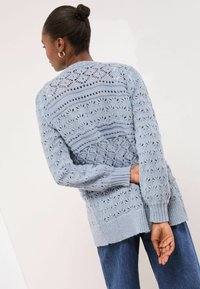 Svetlomodrý pletený kardigán s otvorenými vzormi, rebrovanými manžetami a spodným lemom, a uvoľneným strihom, zladený s modrými džínsami.