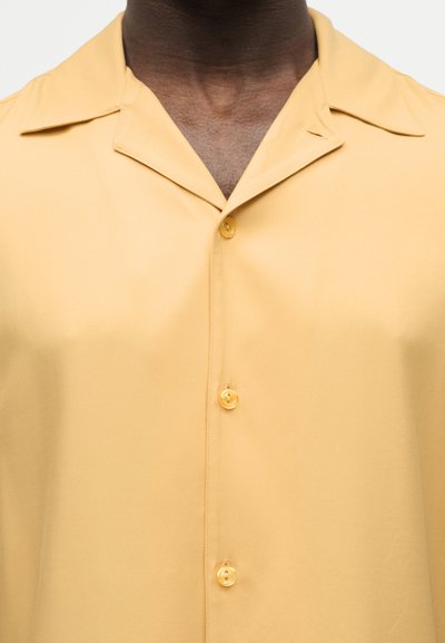 Κοντινό πλάνο ενός ατόμου που φοράει ένα κίτρινο κοντομάνικο πουκάμισο με γιακά τύπου κάμπινγκ, εμφανίζοντας τον θώρακα και το ντεκολτέ.