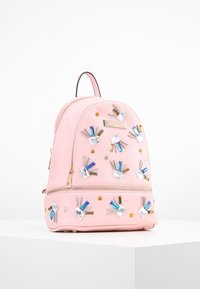 Růžový batoh z umělé kůže s dekorativními krystaly, zakulaceným tvarem a zlatými doplňky. Obsahuje dvojitý zip a nastavitelná ramena.