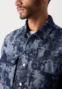Denim jacket in a blue camouflage pattern. Features a collared neckline, snap buttons, and a chest pocket with a logo patch.