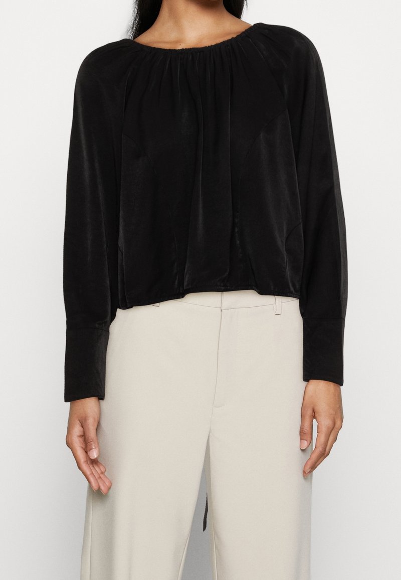 Haut noir à manches longues en tissu lisse et doux avec un col froncé et un drapé subtil ; assorti à un pantalon beige taille haute.