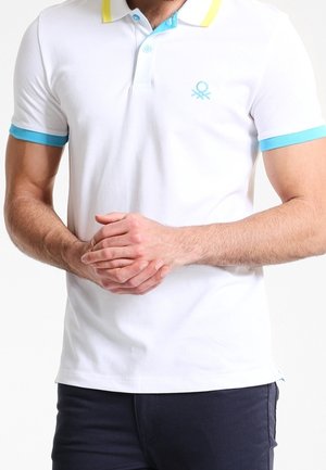 Homme portant un polo blanc avec un col et des poignets jaune et bleu, les mains jointes devant lui, sur un fond uni.