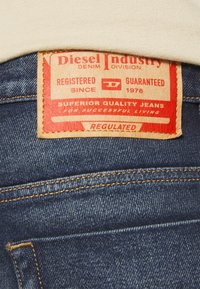 Jeans de mezclilla azul con una etiqueta de cuero marrón. La etiqueta presenta texto rojo y blanco, indicando detalles de marca y calidad. Costuras visibles que acentúan el diseño.