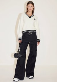 Weißer, flauschiger Pullover mit schwarzen gestreiften Ärmelbündchen und Kragen, kombiniert mit schwarzen Cargo-Hosen. Accessoires umfassen eine runde schwarze Tasche.