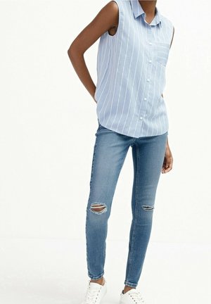 Persona que lleva una camisa de botones sin mangas a rayas azules y blancas, vaqueros pitillo azules rasgados y zapatillas blancas, de pie con las manos detrás de la espalda.