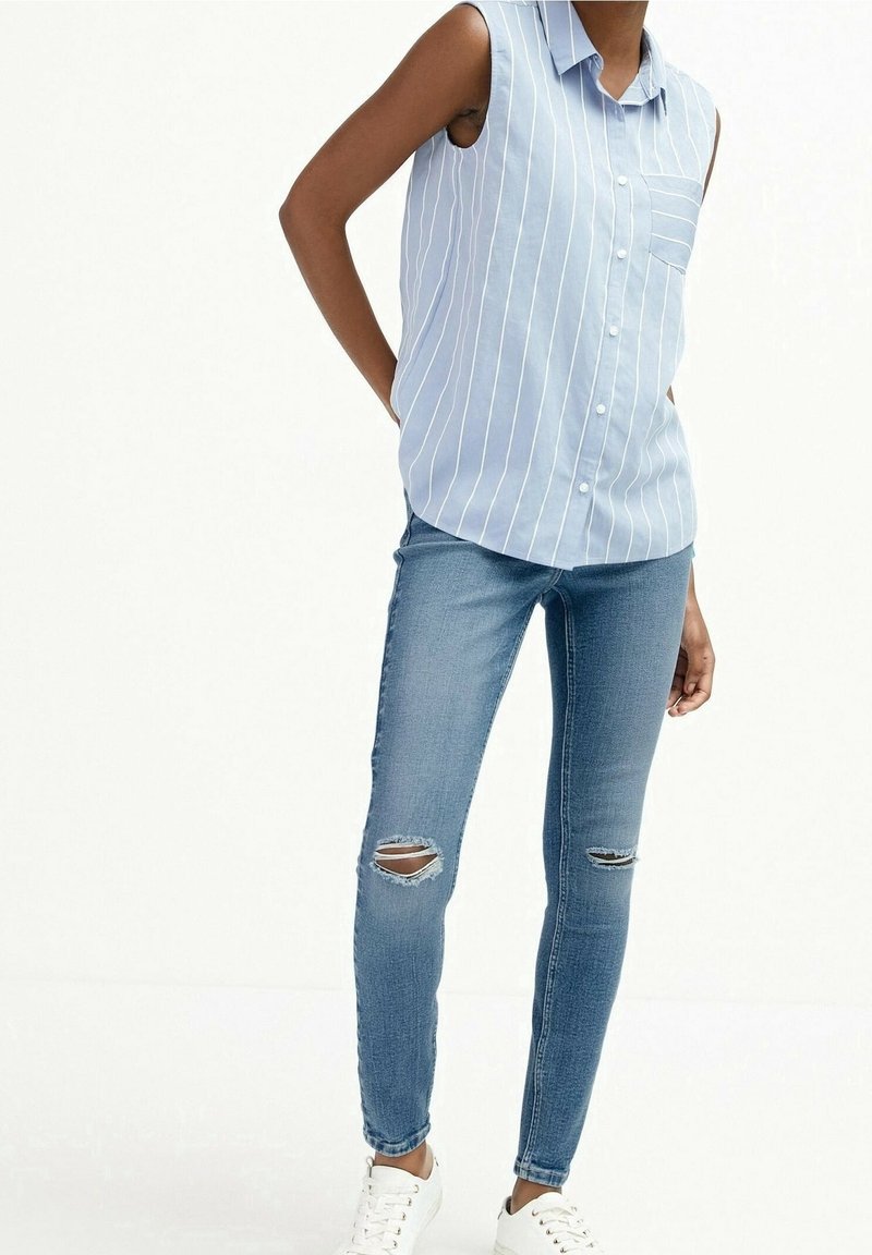 Personne portant une chemise sans manches rayée bleu et blanc à boutons, un jean skinny bleu déchiré et des baskets blanches, debout avec les mains derrière le dos.