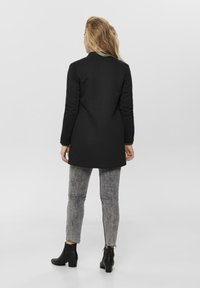 Manteau long noir à coupe droite, associé à un jean gris usé et des bottines noires. Texture lisse, détails minimalistes.