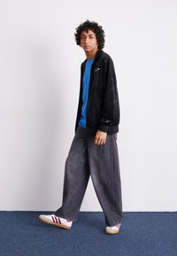 Sort velourjakke over en blå T-shirt, parret med brede grå jeans og hvide sneakers med røde striber. Modellen har krøllet hår.