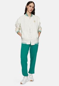 Vit bomberjacka med gröna ränder, tryckknappar och logotyp. Kombinerad med gröna sweatpants och vita sneakers. Slät textur.