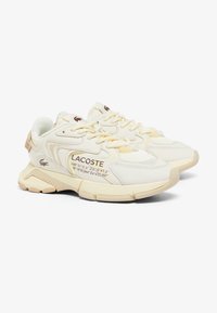 Baskets Lacoste en crème et jaune clair. Matériaux en mesh et synthétiques, col rembourré, accents texturés et détails imprimés sur le côté.