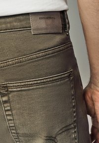Oliwkowe dżinsy o gładkiej fakturze, z tylną kieszenią i brązową metką z napisem "DENIM SUPPLY CO." na pasku.