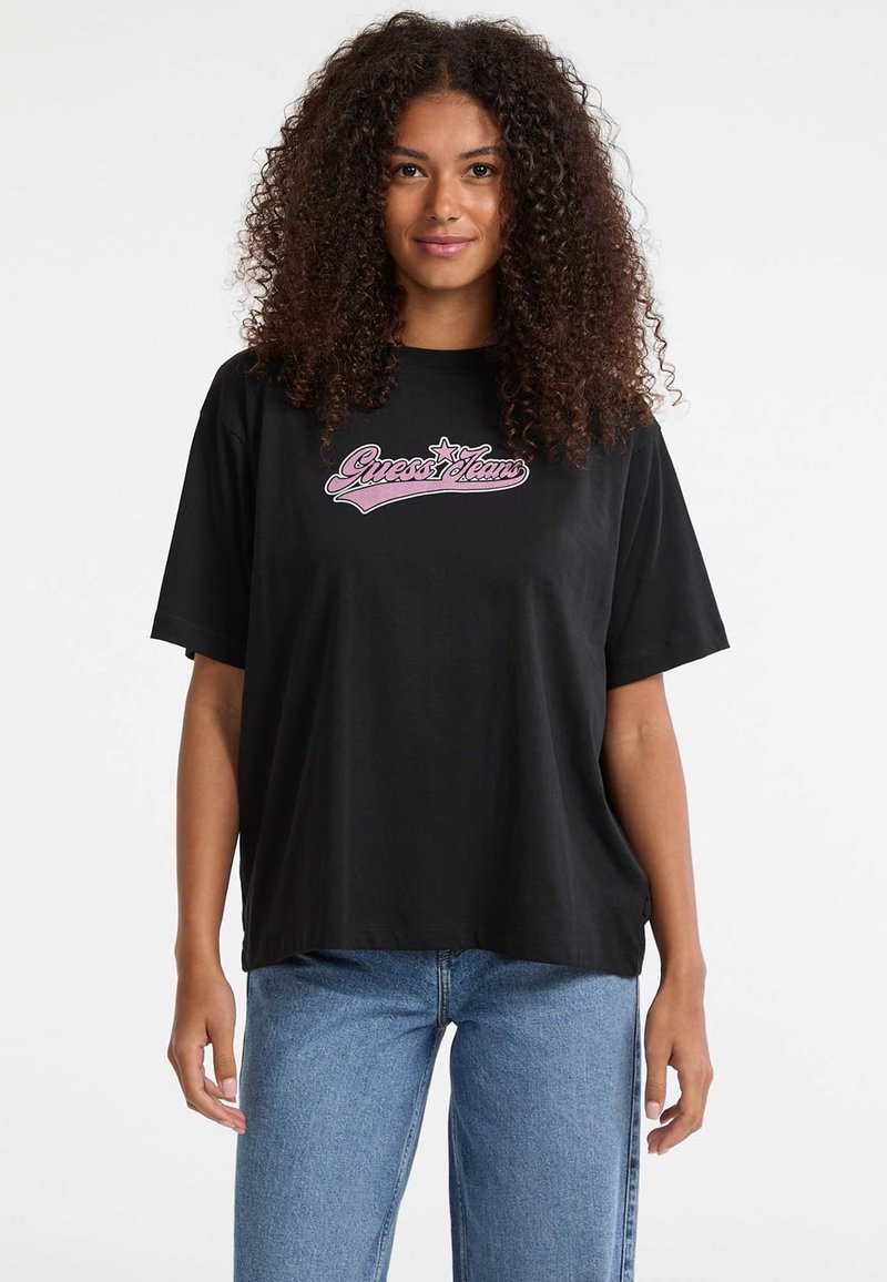 Žena s kovrčavom kosom nosi crni preveliki majicu s ružičastim logotipom "Guess Jeans" i plave traperice, stojeći nasuprot obične bijele pozadine.
