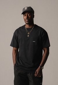 T-shirt en coton noir avec une poche portant "BLKVTS" en broderie blanche. Le modèle porte une casquette grise et des colliers en or superposés. Shorts noirs.