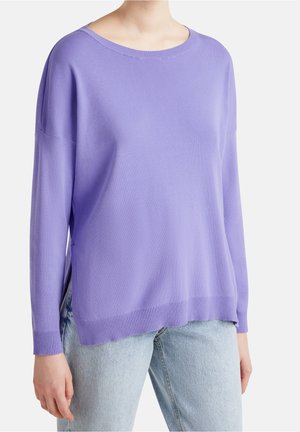 Henkilö pukeutuneena vaaleanlila, pitkähihainen neulepusero, jossa on resorit hihoissa ja helmassa, yhdistettynä vaaleansinisiin farkkuihin.