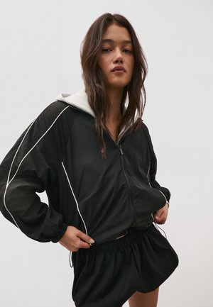 Giovane donna con lunghi capelli scuri che indossa una giacca nera con zip e bordo bianco e pantaloncini neri coordinati su uno sfondo neutro.