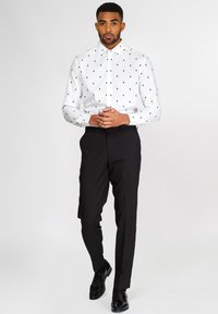Camicia bianca con motivi di alberi verdi, abbinata a pantaloni neri su misura e scarpe eleganti nere lucide.