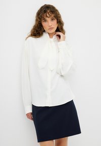 Blouse blanche avec un détail de col noué, à boutons devant et manches longues. Portée avec une jupe bleu marine foncé, présentant une texture de tissu lisse.