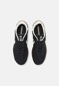 Baskets en daim noires avec des marques et accents blancs, présentant un design à lacets, une semelle intérieure rembourrée et une semelle extérieure en caoutchouc brun.