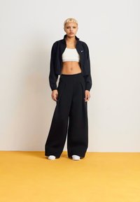 Sort cropped jakke over en hvid crop top, parret med brede sorte bukser. Modellen står på en gul baggrund og har hvide sneakers på.