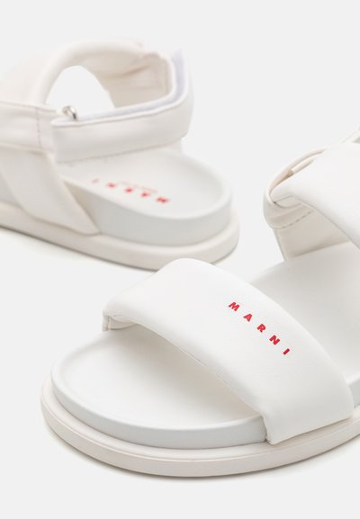 Sandalias blancas con correas anchas, con una textura suave y un logo en rojo. La forma es abierta en la parte delantera con una suela acolchada y cierre ajustable.