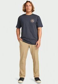 Hombre con cabello rizado hasta los hombros, vistiendo una camiseta gráfica azul oscura, pantalones beige y zapatillas negras, de pie con una mano en el bolsillo.