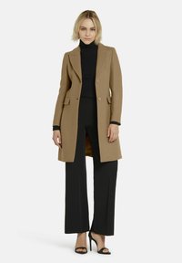 Camelkleurige op maat gemaakte jas met dubbele knopen, notch-revers en zijzakken, gedragen over een zwarte coltrui en zwarte broek.