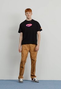 Črna majica s kratkimi rokavi Nike z vijoličnim logotipom, v kombinaciji s khaki cargo hlačami in črno-belimi supergami z rdečimi poudarki.