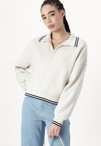 Hanorac cu guler din amestec de bumbac de culoare alb-îngălbenită. Prezintă dungi contrastante de culoare bleumarin pe guler și manșete, având un design scurt.