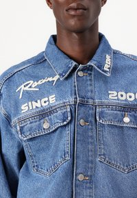 Chaqueta de denim azul con texto bordado "Racing", "SINCE" y "2000" con botones plateados y dos bolsillos frontales. Diseño de collar estructurado.