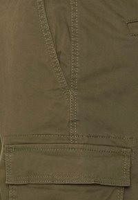 Pantalons cargo vert olive avec des coutures visibles, featuring une poche latérale et une poche plate à l'avant. Le matériau semble durable et texturé.