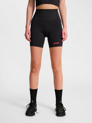 Vrouw draagt zwarte high-waisted sportshorts met rood "hummel" logo, zwarte sokken en zwarte hardloopschoenen, staand tegen een effen achtergrond.