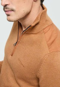 Pull en maille à demi-fermeture éclair de couleur marron chaud, avec col côtelé et panneaux d'épaule contrastants en beige. Texture subtile et détails de marque minimaux.