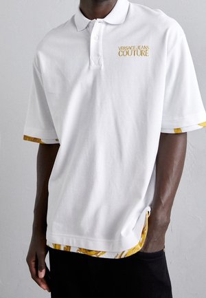 Hvid polo-shirt lavet af struktureret stof, med et guldbroderet logo og livligt gult mønsterbånd på ærmerne og nederkanten.