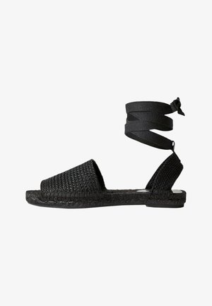 Sandales noires tressées avec une lanière de cheville. Dessus texturé et semelle plate en jute. Design simple sans matériel visible.