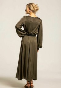 Olivgrön klänning med långa ärmar, en åtsittande svart midja och ett blixtlås i ryggen, med en svävande kjol som når anklarna.