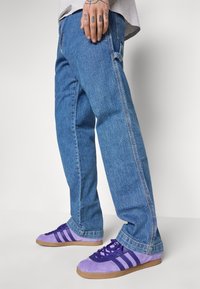 Blugi largi, de culoare albastră, cu un finisaj decolorat, din material denim, asortați cu teniși din suede mov, cu trei dungi și tălpi din cauciuc maro.