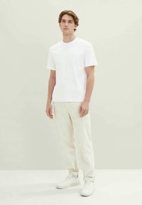 T-shirt branca de algodão com mangas curtas, combinada com calças bejes claras e sapatilhas brancas; design minimalista contra um fundo neutro.