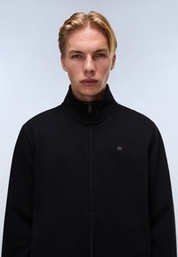 Černá zipová bunda s vysokým límcem. Materiál se zdá hladký, s jemnou strukturou. Obsahuje malé logo na levém hrudníku.
