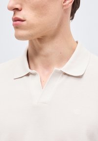 Νέος άνδρας που φοράει ένα λευκό πολο με υφή και μικρό λογότυπο "ck" στο στήθος, κοιτάζοντας προς τα αριστερά σε απλό φόντο.