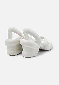 White rubber sandals with a unique sculptural design, featuring cut-out shapes and a curved chunky heel. Smooth texture and minimal accents.