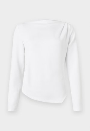 Blouse blanche à manches longues avec encolure bateau et ourlet légèrement asymétrique, présentée sur un fond clair uni.