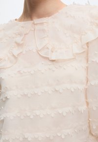 Blouse transparente crème avec des bandes horizontales d'appliques florales et un col à volants.