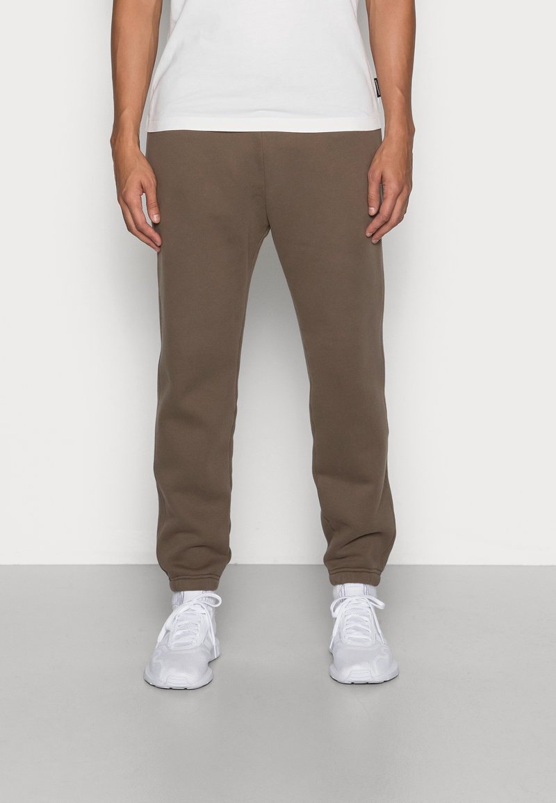 Persoon in een bruine sweats broek, witte sneakers en een wit T-shirt die tegen een simpele witte muur en een grijze vloer staat.