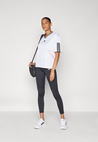 Balts sporta krekls ar melnām svītrām, rozā akcentu, apvienots ar tumši pelēkiem legingiem un baltām sporta apaviem. Modelis tur melnu somu.